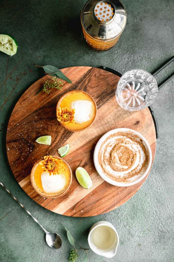 Two margaritas on a circular wooden tray garnished with yellow mums. Greenery, lime wedges and a small plate of pumpkin spice and salt mix on the tray. A cocktail shaker and cocktail spoon to the side.