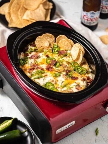 Jalapeno Popper Dip in a red crock pot with a black insert. A couple bottles of beer, tortilla chips and whole jalapenos on black plates to the side of the crock pot.