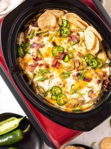 A red crockpot with a black insert filled with cooked jalapeno popper dip and topped with bacon, green onion, crispy jalapenos and round tortilla chips. Black plates of jalapenos and tortilla chips to the side.