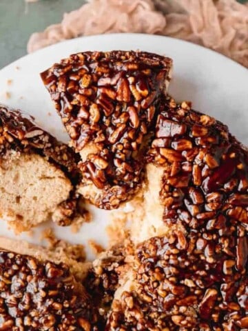 Moist vanilla cake topped with a gooey pecan glaze, sliced into pieces on top of a white circular tray.