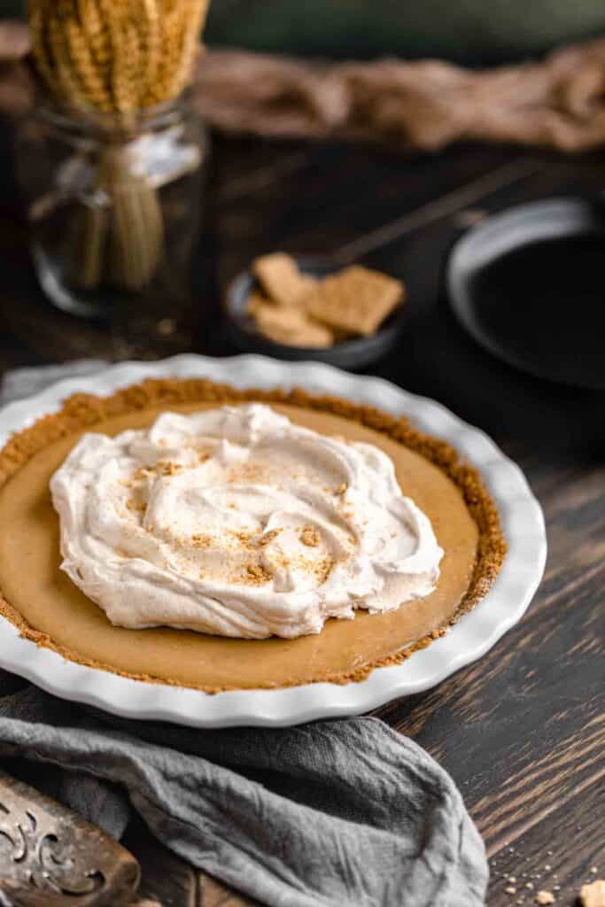 Cinnamon Butterscotch Pie with whipped topping in a white pie pan on a dark wood table. A blue linen towel with a black plate of graham crackers behind it. 