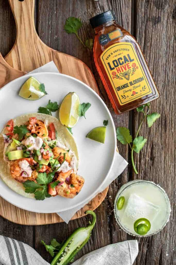 A shrimp taco on a white plate with a bottle of Local Hive Honey, margarita and a jalapeno next to it.