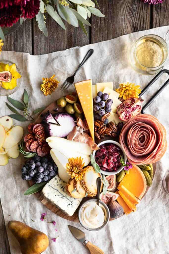 An Easy Thanksgiving Charcuterie Board with meats, cheeses, fruits, nuts, spreads and crackers on a linen with flowers and fruit surrounding it.