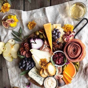 A beautifully arranged board with meats, cheeses, cranberry sauce, pumpkin goat cheese whip, crackers and fruits. Sitting on top of a linen with flowers and a glass of white wine next to it along with a whole pear and a few thin apple slices.