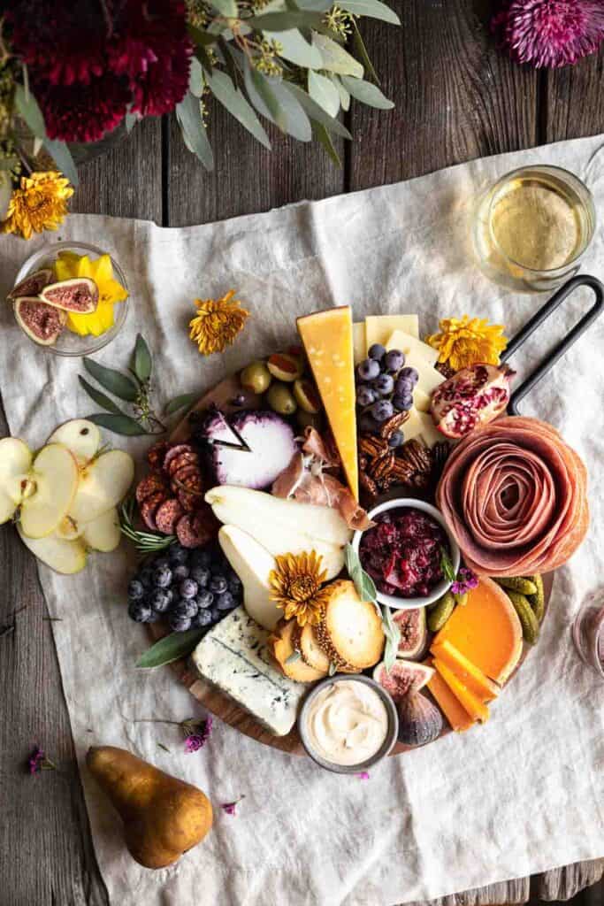 A beautifully arranged board with meats, cheeses, cranberry sauce, pumpkin goat cheese whip, crackers and fruits. Sitting on top of a linen with flowers and a glass of white wine next to it along with a whole pear and a few thin apple slices.