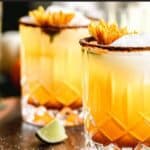 Two pumpkin spice margaritas on a circular wooden tray with large ice cubes, yellow mums and lime wedges near a window.
