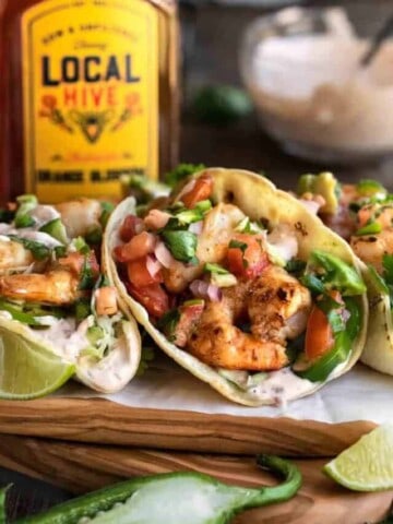 Tacos de Camaron on a wooden cutting board with white parchment paper underneath. A bottle of Local Hive Honey and bowl of chipotle sour cream behind the tacos.