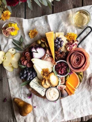 A beautifully arranged board with meats, cheeses, cranberry sauce, pumpkin goat cheese whip, crackers and fruits. Sitting on top of a linen with flowers and a glass of white wine next to it along with a whole pear and a few thin apple slices.