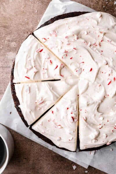 Circular chocolate peppermint cake with three slices cut but not removed. Sitting on white parchment paper on a brown surface and topped with crushed candy cane.