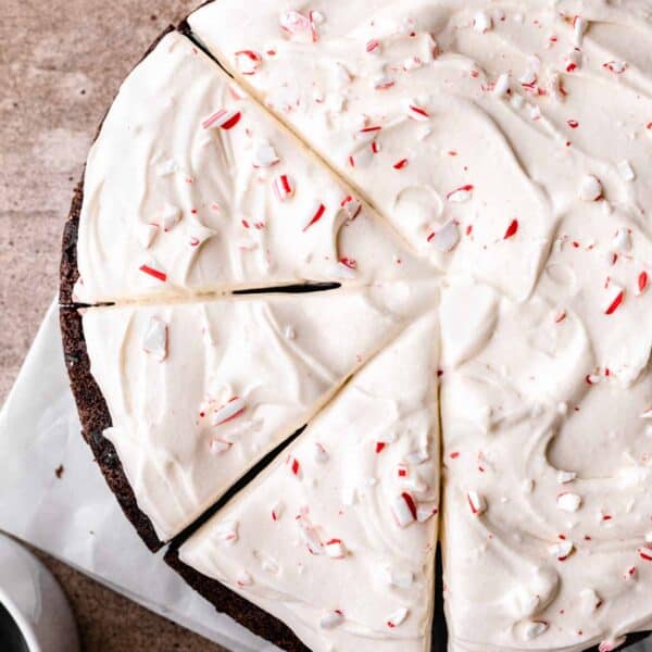 Circular chocolate peppermint cake with three slices cut but not removed. Sitting on white parchment paper on a brown surface and topped with crushed candy cane.