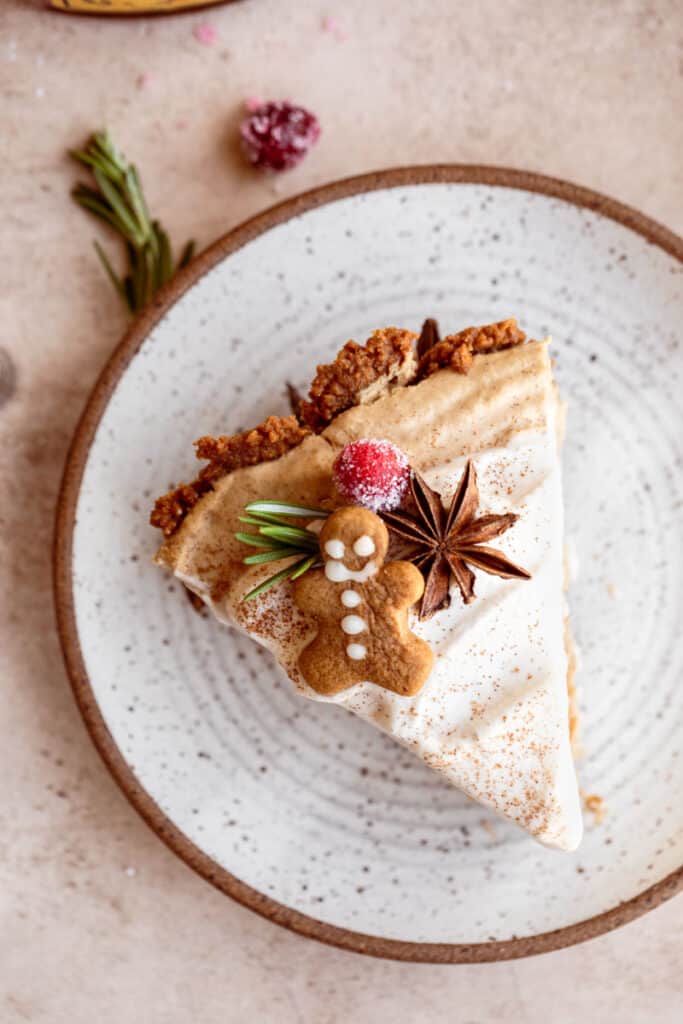 A slice of pie topped with a tiny gingerbread man, star anise, sugared cranberry and rosemary on top of a white speckled plate.