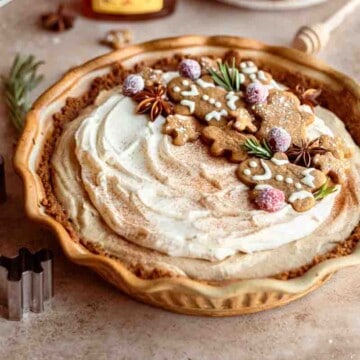 Gingerbread Cream Pie with gingerbread cookies, sugared cranberries, star anise and rosemary on top to garnish. A bottle of Local Hive Honey and plate of cookies behind the brown pie pan.
