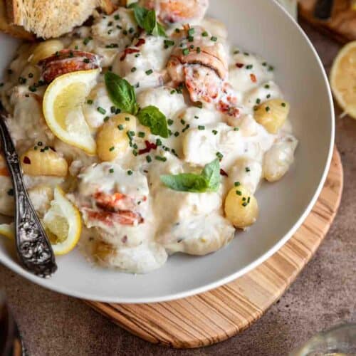 Gnocchi with lobster and parmesan cream sauce in a bowl topped with basil, chives and a few pieces of toasted baguette. A glass of white wine and lemon sitting to the side of the bowl.