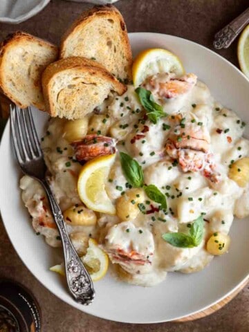 Lobster Gnocchi in a white bowl topped with fresh basil, chives, lemon wedges and a few pieces of toasted baguette. A fork in the bowl.