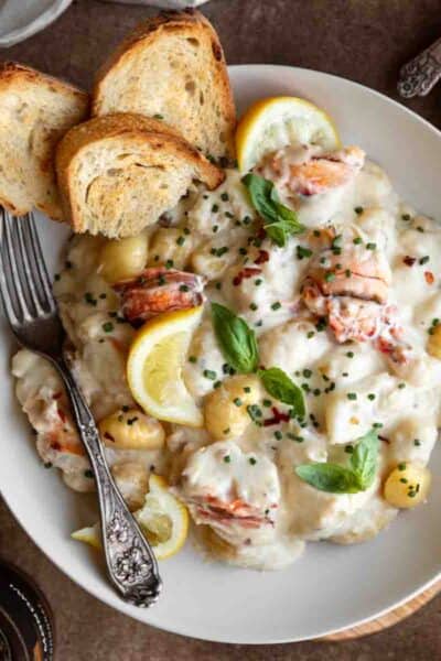 Lobster Gnocchi in a white bowl topped with fresh basil, chives, lemon wedges and a few pieces of toasted baguette. A fork in the bowl.