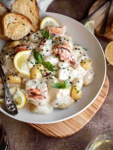 Gnocchi with lobster and parmesan cream sauce in a bowl topped with basil, chives and a few pieces of toasted baguette. A glass of white wine and lemon sitting to the side of the bowl.