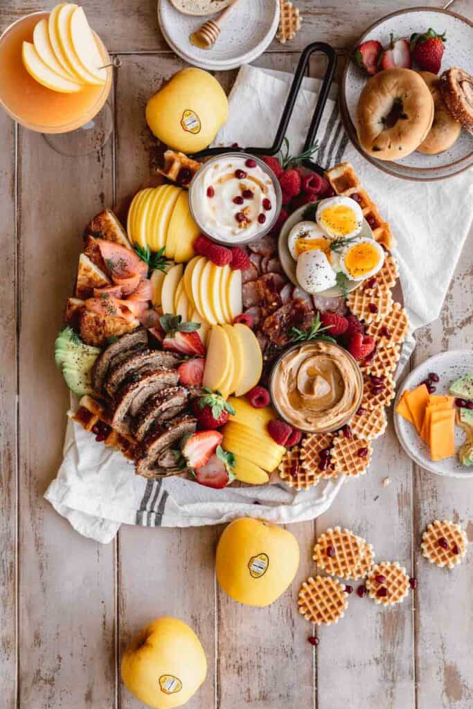 Breakfast charcuterie board filled with apples, peanut butter, yogurt, bacon, sausage, eggs, waffles, fresh fruit, smoked salmon and assorted bread.