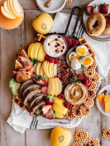 Breakfast charcuterie board filled with apples, peanut butter, yogurt, bacon, sausage, eggs, waffles, fresh fruit, smoked salmon and assorted bread.