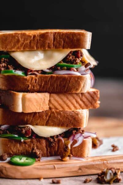 Texas Toast Sloppy Joes stacked on top of each other on a wooden cutting board with melting cheese coming over the edge of the sandwich.