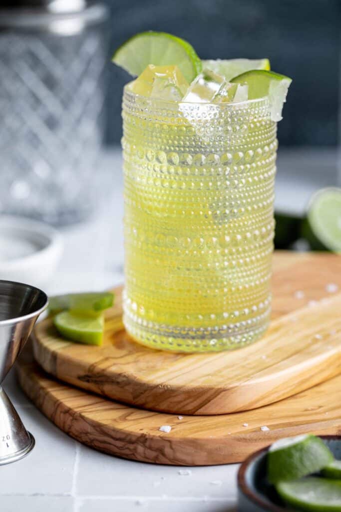 A green tea shot cocktail in a texture clear glass sitting on top of 2 wooden cutting boards with lime wedges nearby.
