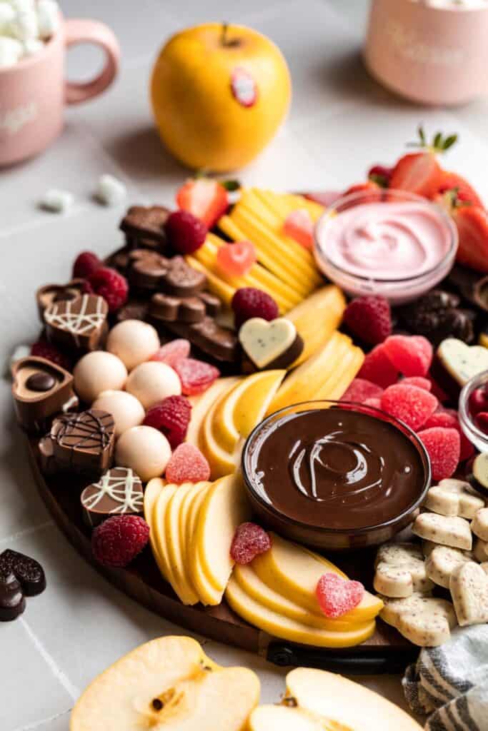 A bowl of melted dark chocolate on a board with fruits, candles and pink mugs filled with marshmallows. 