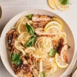 A bowl of lemon chicken pasta with lemon chicken and creamy sauce over the top. Fresh basil leaves, lemon slices and a serving spoon next to the bowl.