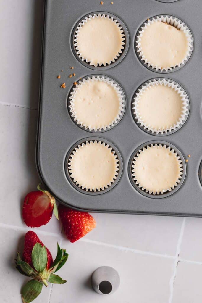 Mini cheesecakes in a pan after being baked with a few strawberries lying to the side. 
