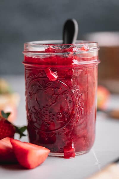 A small mason jar filled with strawberry compote that has a spoon coming out of the top and strawberries laying to the side.