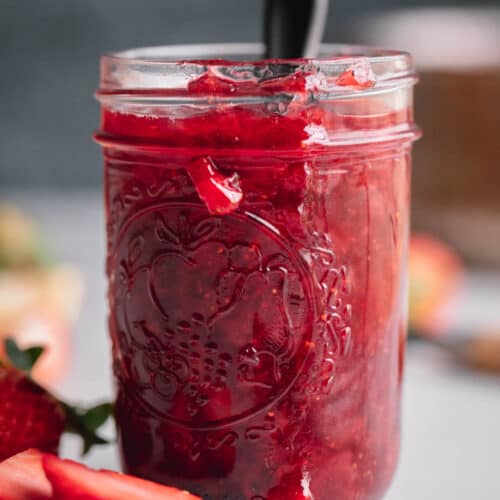 A small mason jar filled with strawberry compote that has a spoon coming out of the top and strawberries laying to the side.
