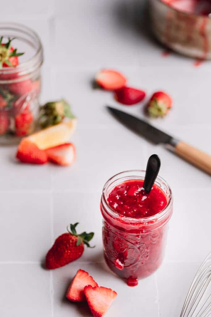 A jar of strawberry compote, a small knife, strawberries and lemon wedges.