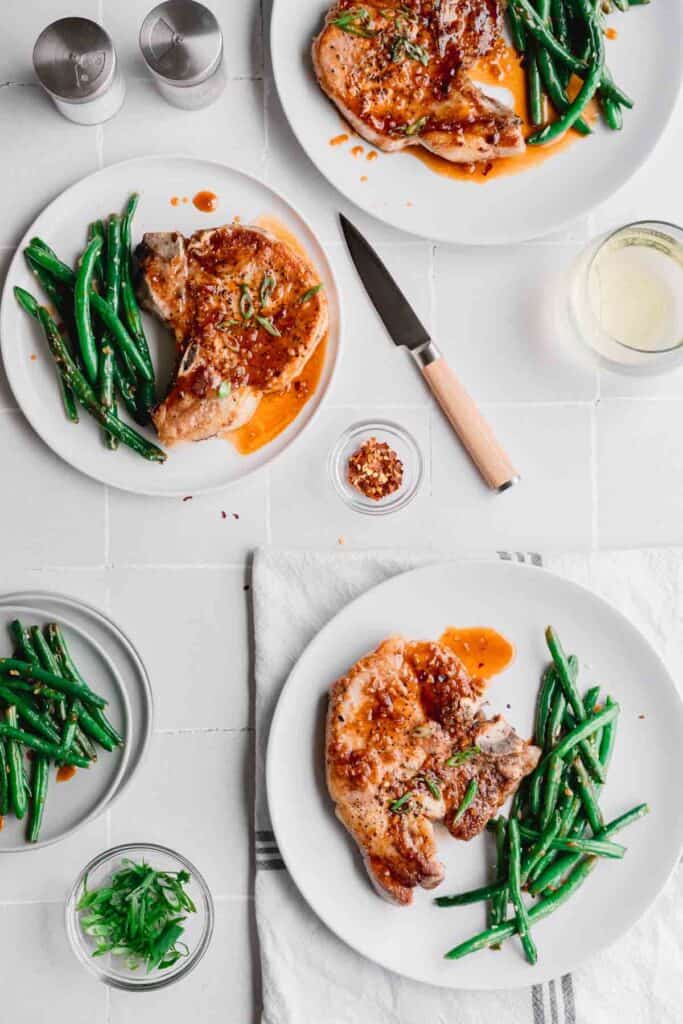 Honey Garlic Pork Chops on three separate plates with green beans, sauce and green onions. A glass of white wine, small knife, salt and pepper shakers to the side. 