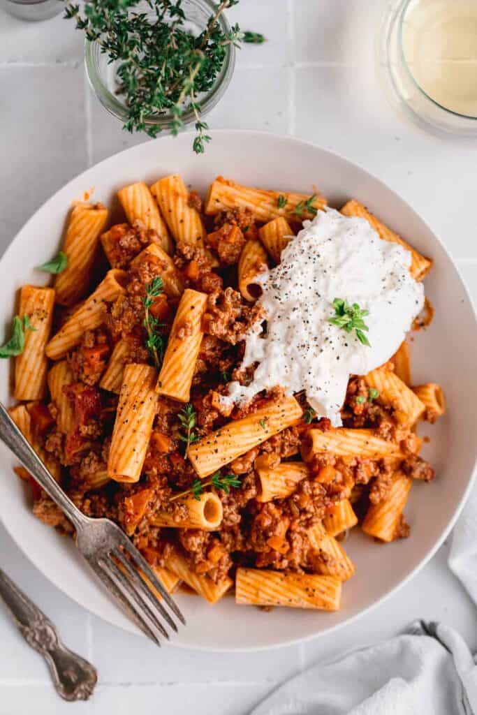 Hearty rigatoni and rich meaty tomato sauce in a white bowl with burrata cheese on top.