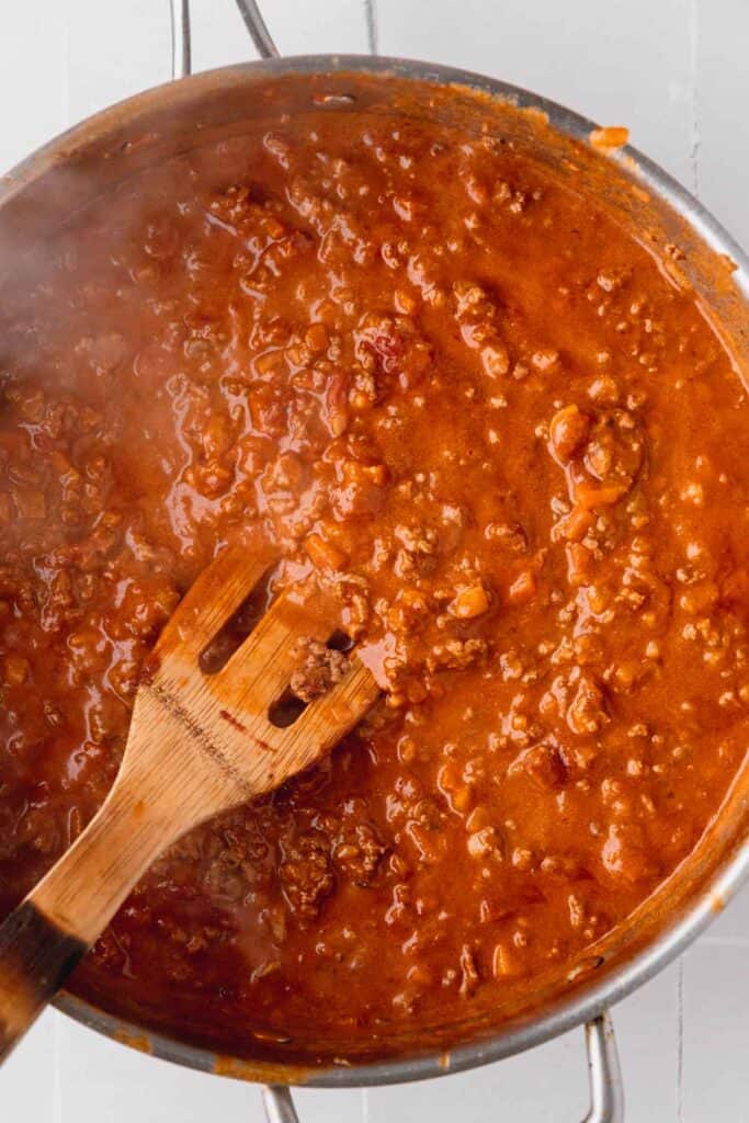 Bolognese sauce simmering in a large skillet with a wooden spatula. 