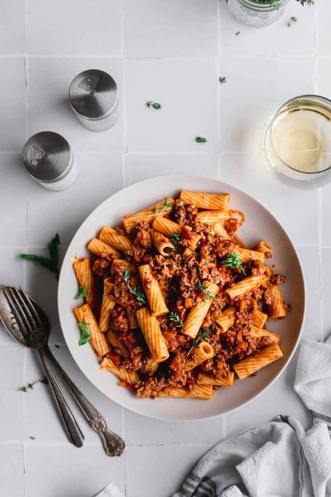A bowl of freshly cooked rigatoni tossed in a bolognese sauce with silverware, wine, salt and pepper to the side.
