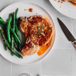 A honey garlic pork chop on a white plate smothered in sauce with a side of green beans. Another plate and a paring knife to the side.