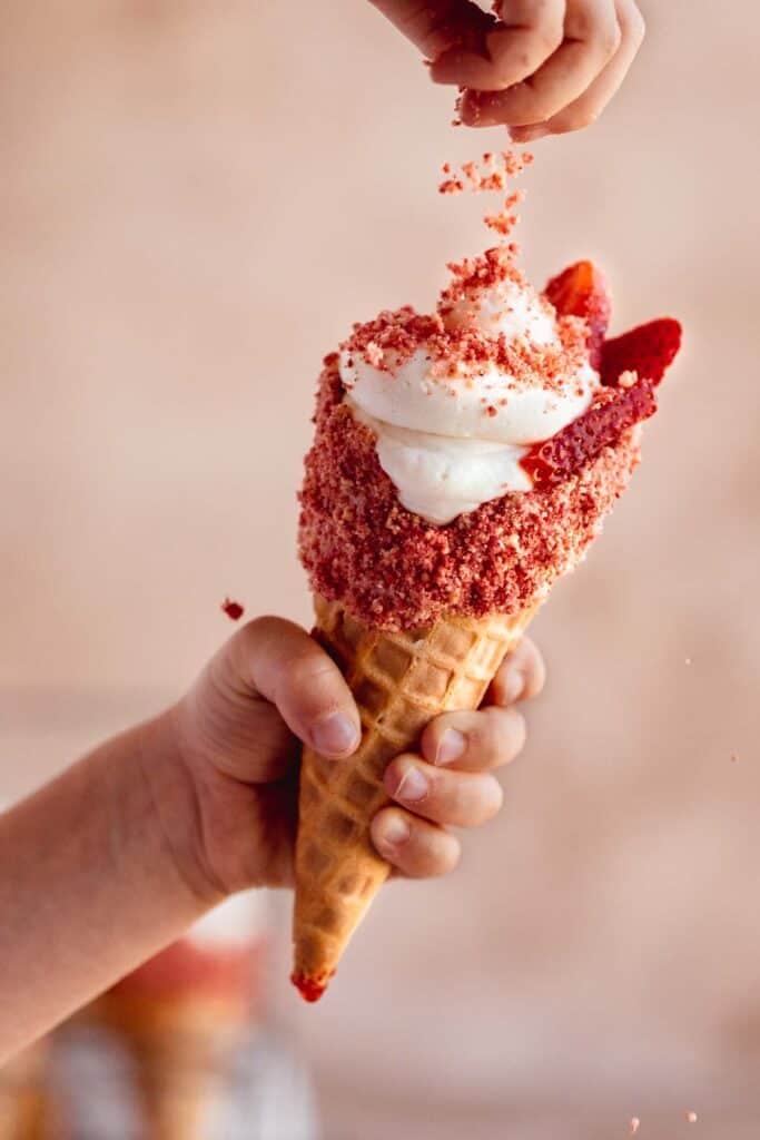 Strawberry crunch topping being sprinkled onto no bake cheesecake filling in a waffle cone.