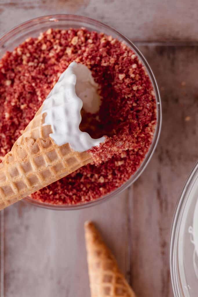 A waffle cone with melted vanilla wafer being dipped into strawberry crunch topping.