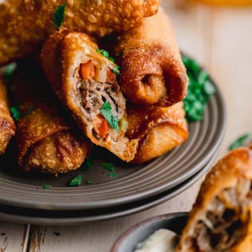 Philly cheesesteak egg rolls stacked up on a plate. One egg roll cut open and dipping sauce to the side.