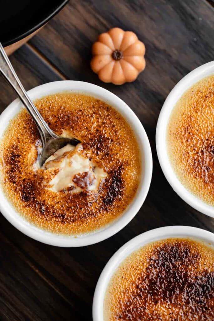 Pumpkin creme brulee in ramekins with a spoon broke through the sugar topping and a pumpkin to the side.