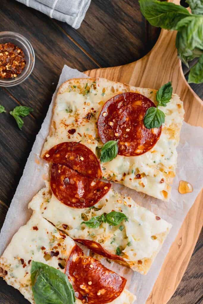 Garlic bread pizza on a wooden cutting board with pepperoni and fresh basil.