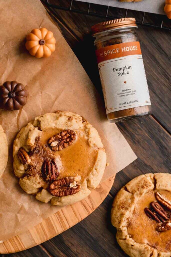 Pumpkin cheesecake cookies topped with brown sugar and pecans. A bottle of pumpkin spice seasoning and candy pumpkins to the side.