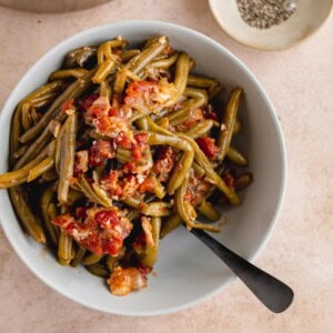 A bowl of green beans stewed with bacon, tomato and onion with a spoon in it.