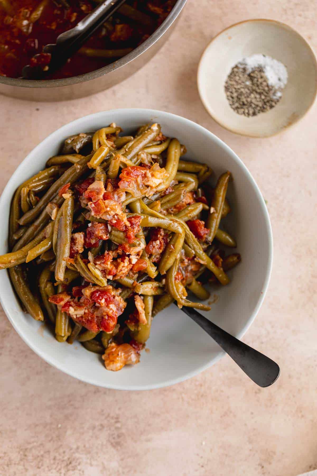 Green Beans With Bacon in a bowl with a spoon and some seasoning to the side. 