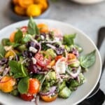 Mediterranean Cucumber Salad with tomatoes, red onion, olives, fresh herbs and red wine vinaigrette in a bowl with spoons to the side.