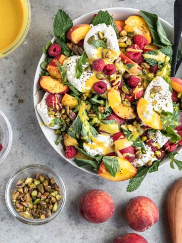 Peach Burrata Salad in a bowl with honey mustard dressing, spoons and ingredients around the outside of the bowl.