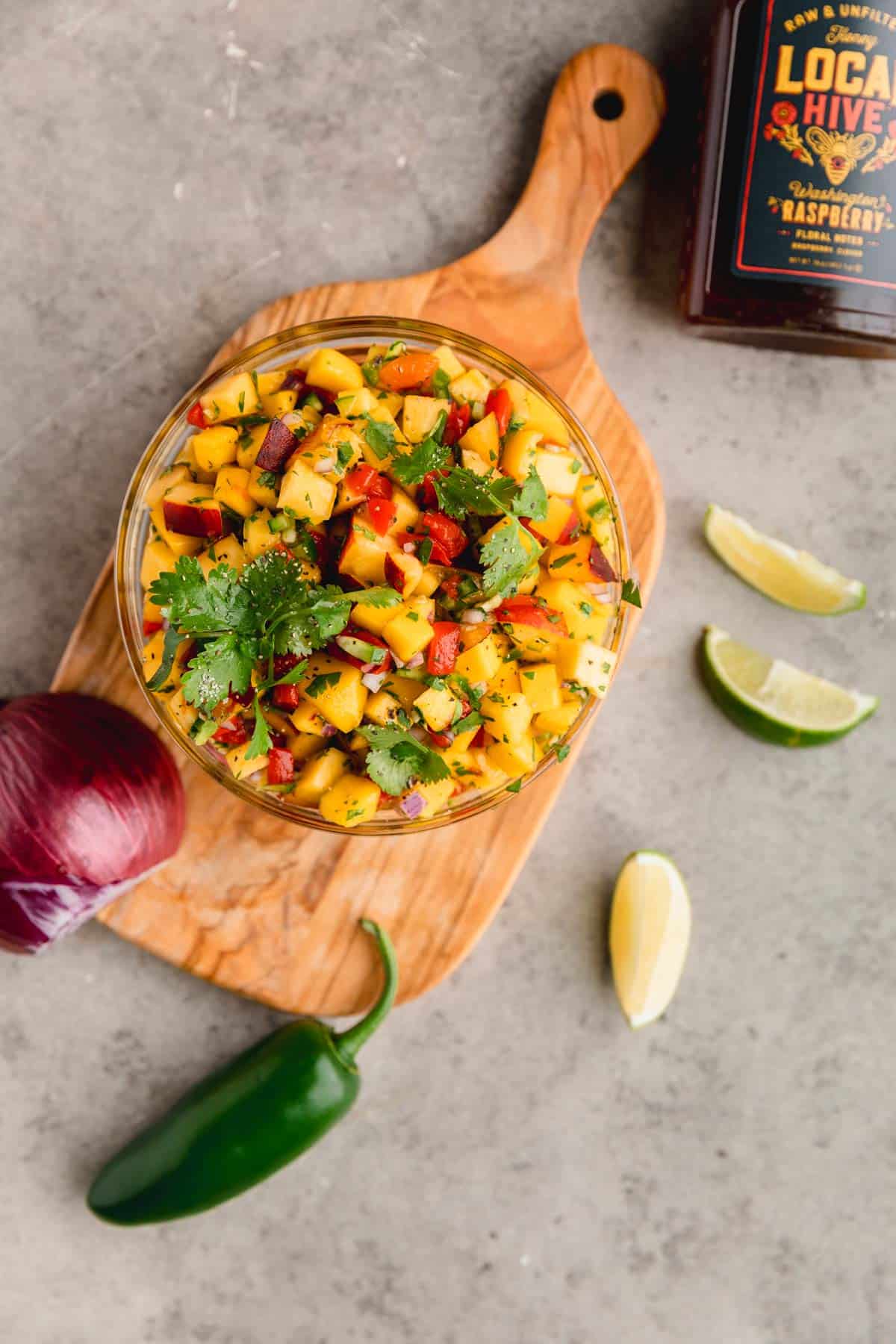 Peach mango salsa in a bowl on a cutting board with lime wedges, jalapeno and onion around it. 