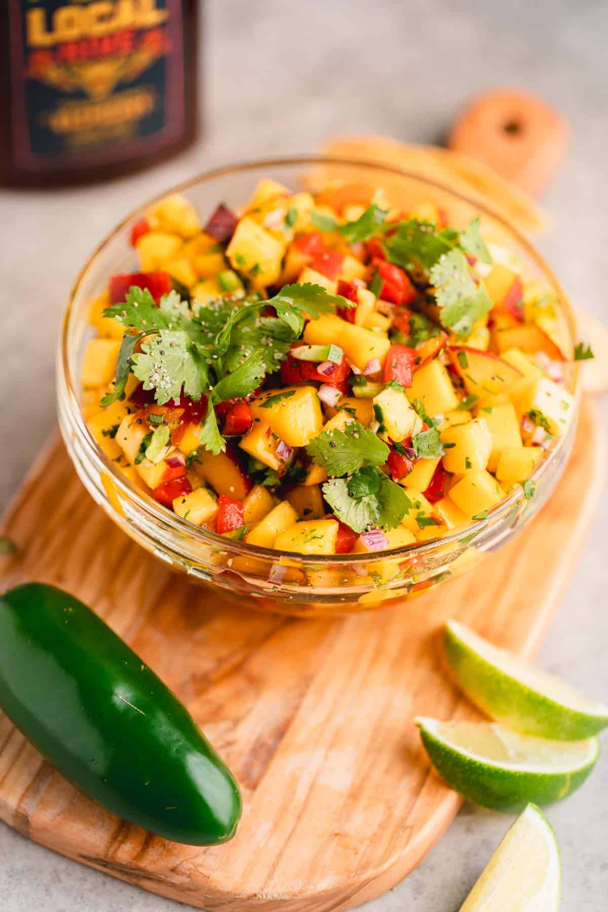 Peach Mango Salsa in a bowl on a cutting board with lime slices and a jalapeno near by. 