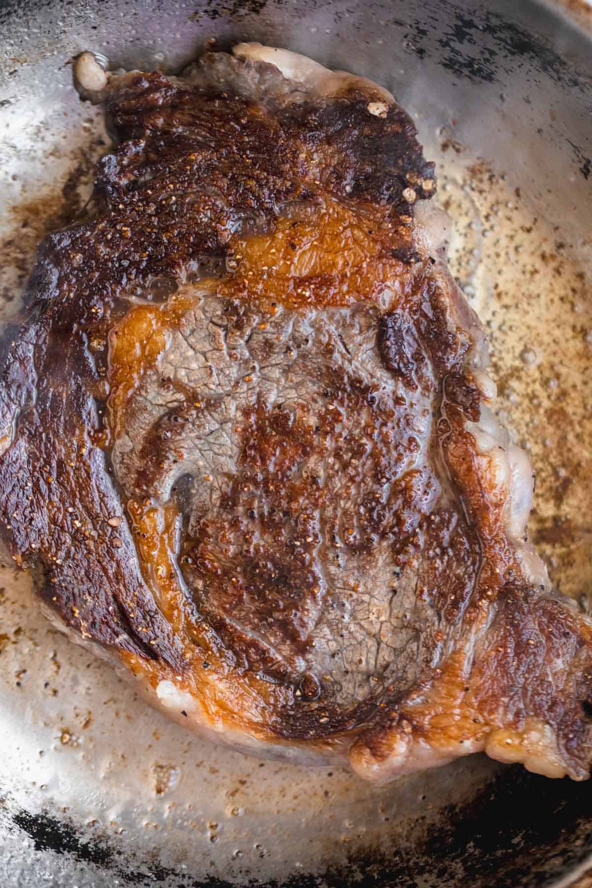 Seasoned ribeye steak cooking in a pan.