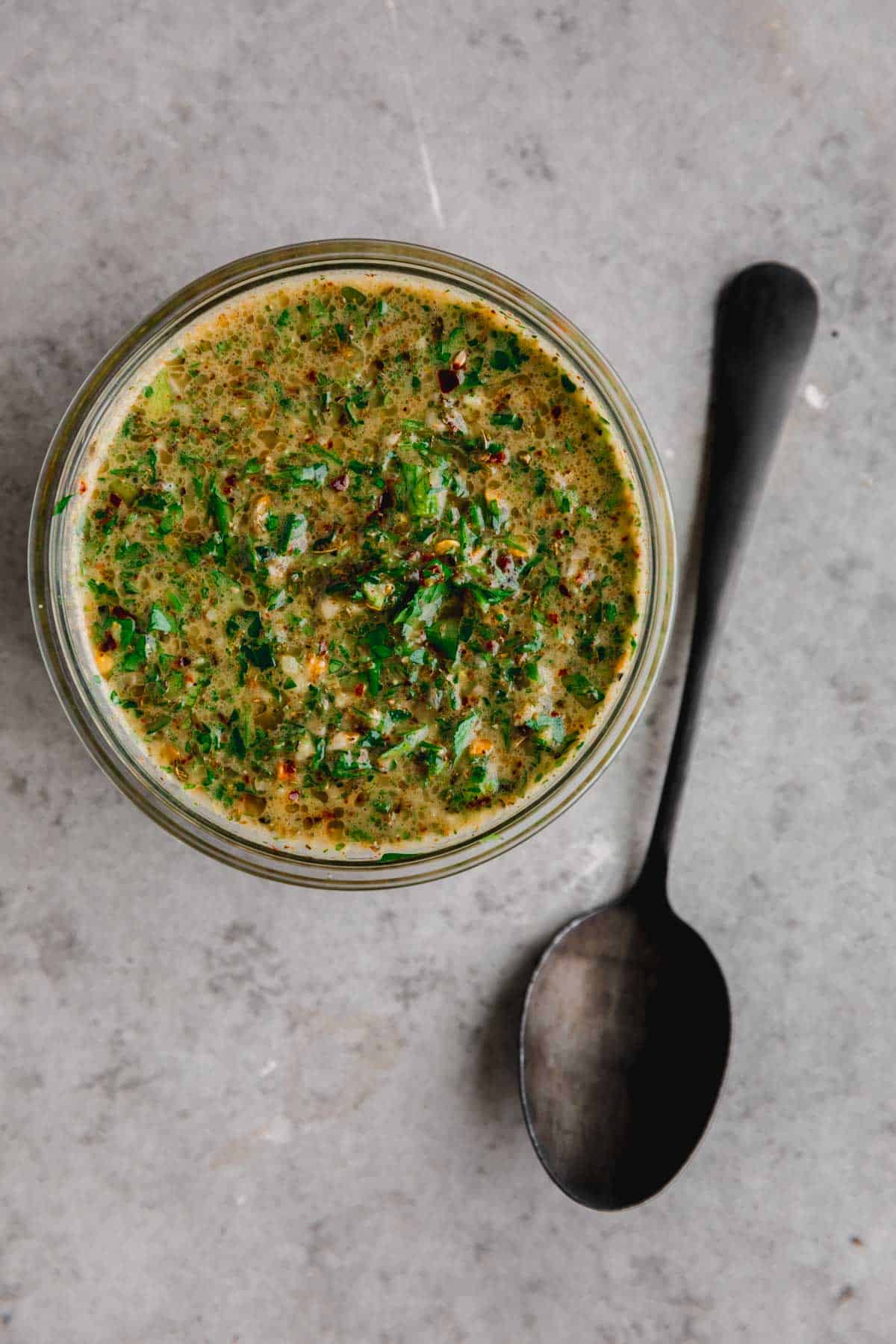 Chimichurri in a small glass dish with a spoon lying to the side.