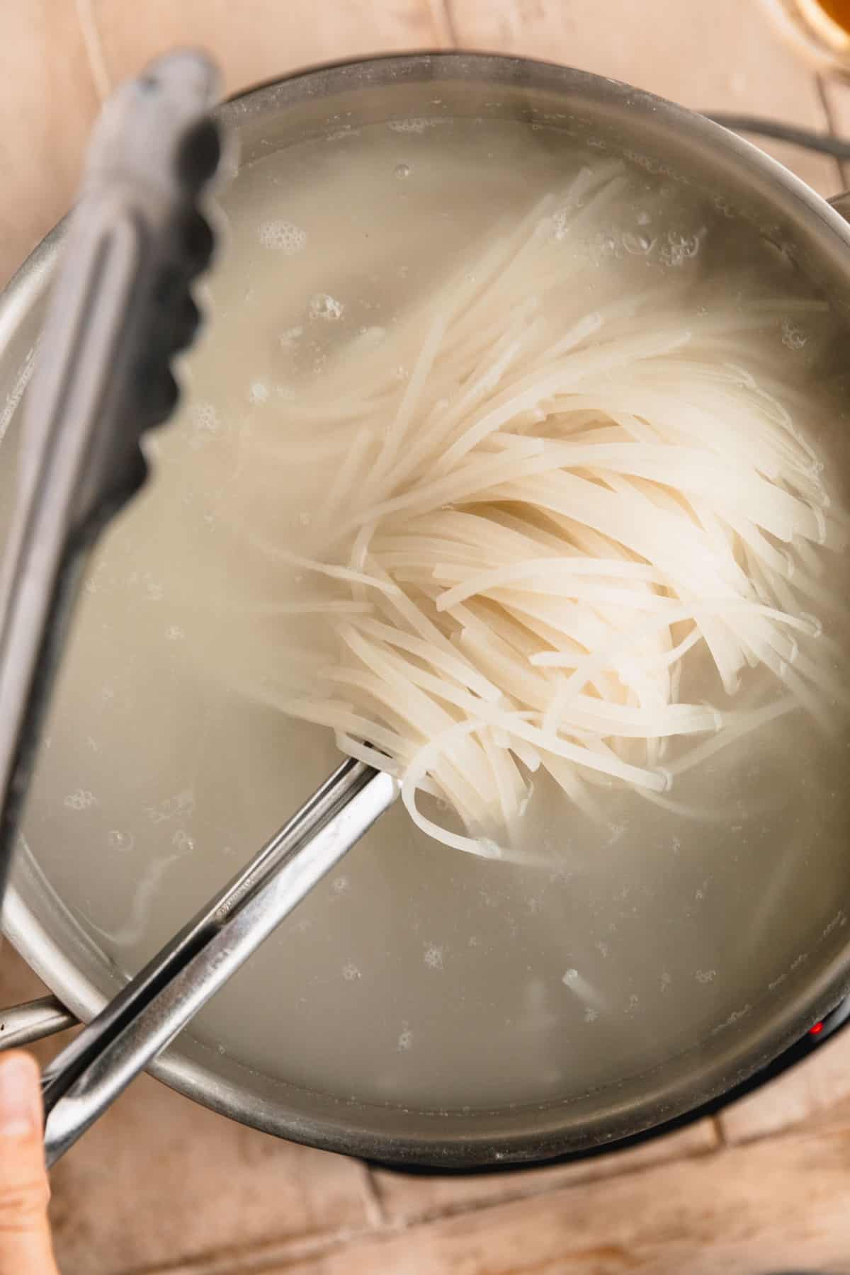 Rice noodles being cooked in a pot of water with tongs lifting the noodles up.